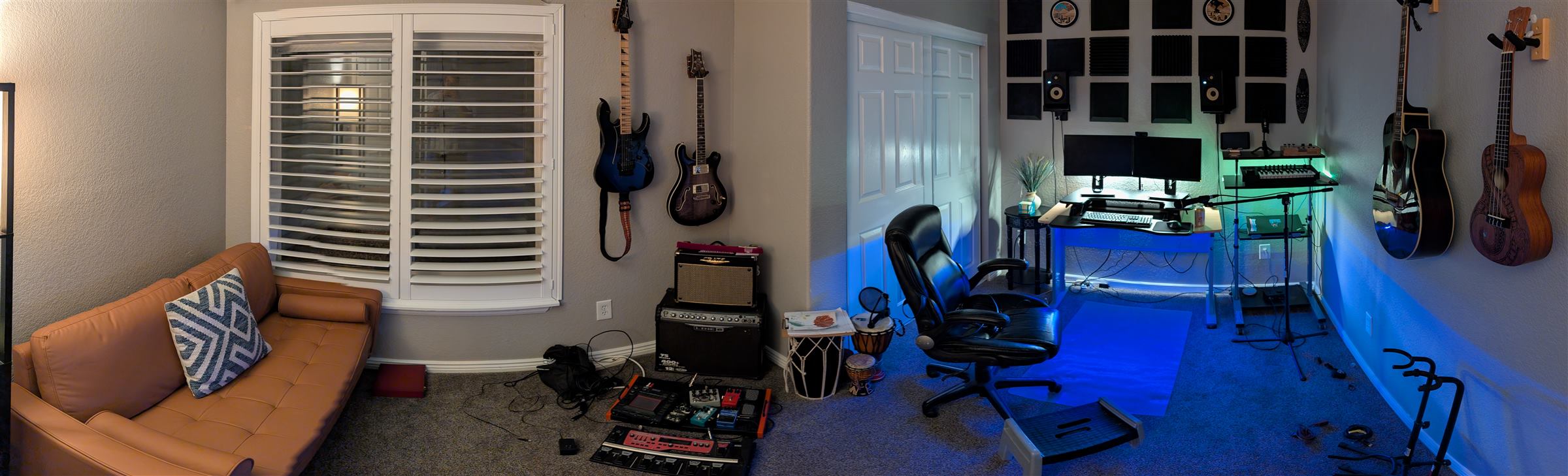 Panoramic view of the Entropic Labs studio room: an orange leather couch beneath a shuttered window, two electric guitars mounted on the left wall above an amp stack and pedalboard, and to the right a desk with dual monitors, acoustic foam panels, and a ukulele and acoustic guitar on the wall, all lit with blue and green LED lighting.